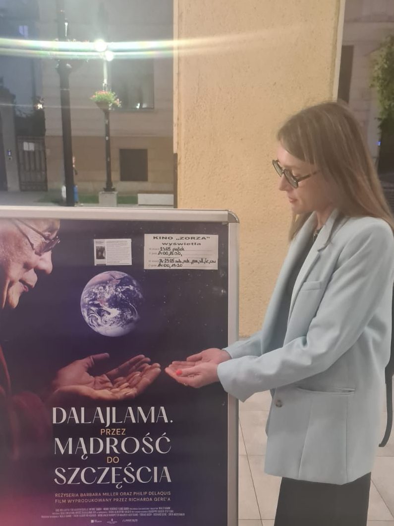 Zdjęcie przed kinem Zorza w Rzeszowie. Kobieta stoi przy plakacie z reklamą filmu Dalajlama. Przez Mądrość do szczęścia.